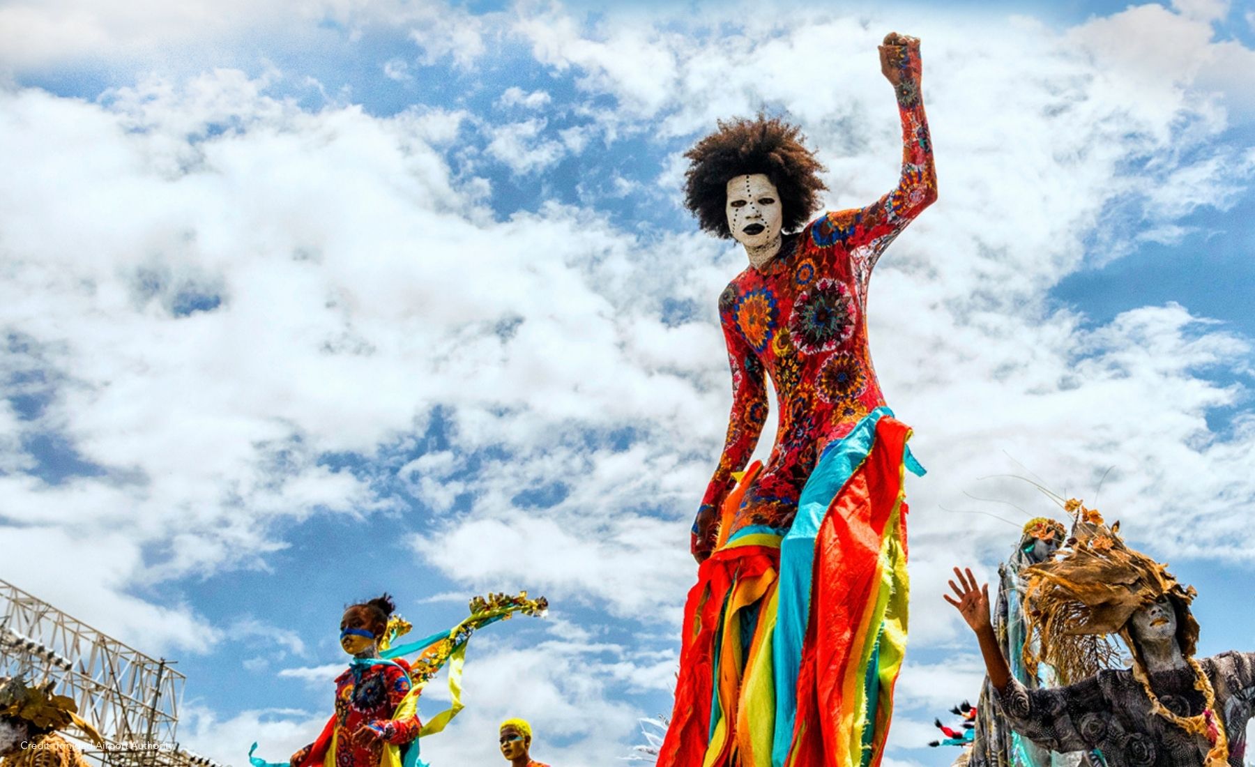 Carnival performer in a colorful costume on stilts during Trinidad Carnival under a bright blue sky.h